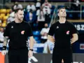 Dwóch sędziów w czarnych koszulkach z logo Hummel i emblematem związku sportowego, gotowych do rozpoczęcia meczu ligi piłki ręcznej.