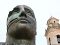Brązowa, monumentalna rzeźba głowy z widocznym fragmentem dzwonnicy kościelnej w tle.
