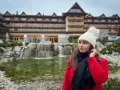 Kobieta w czerwonym płaszczu i białej czapce przed hotelem Bania. W tle wodospad i staw. Wiele pozytywnych hotel Bania opinie potwierdza ten widok.