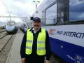 Uśmiechnięty rewident taboru kolejowego w kamizelce odblaskowej stoi obok pociągu PKP Intercity.