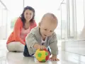 Maluch ćwiczy motorykę dużą, raczkując po podłodze i bawiąc się kolorową piłeczką. Uśmiechnięta mama obserwuje jego postępy.