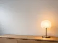En stilren lila lampa lyser upp en träbyrå mot en texturerad vägg.