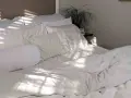 Cama blanca con luz solar filtrada. Aprende como lavar un edredón de plumas para mantenerlo esponjoso.