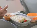 Ręka trzyma telefon nad tacą z jedzeniem: kanapkami, deserem i napojem. Czy w bagażu podręcznym można mieć jedzenie? Tak, jak widać na zdjęciu.