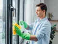 Mężczyzna w zielonych rękawiczkach czyści okna plastikowe, używając sprayu i żółtej ściereczki. Dbałość o detale to klucz do konserwacji okien.