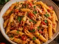 Penne z tuńczykiem i pomidorami, posypane natką pietruszki. Obok puszka tuńczyka, pomidory i czosnek.
