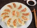 Chrupiące pierożki gyoza ułożone na talerzu, z chrupiącą, złotą panierką. Obok miseczka sosu sojowego i pałeczki.