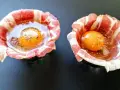 Due uova crude con tuorlo brillante, avvolte in strisce di bacon croccante, pronte per essere cotte.