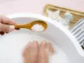 A hand holds a wooden spoon with coarse salt, about to add it to a white basin where feet are soaking. This is a relaxing foot soak with baking soda.
