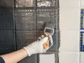 Ręka w białej rękawiczce maluje pędzlem szare płytki. Widać postęp prac, część płytek jest już pomalowana.