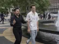 Il cast di And Just Like That, Mario Cantone e Billy Eichner passeggiano ridendo vicino a una fontana in una giornata nuvolosa.