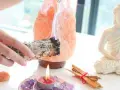 Manos sostienen un manojo de salvia humeante, junto a una lámpara de sal rosa, una vela encendida y una estatua de Buda. Esencial para la limpieza energética de casa.
