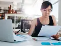 Junge Frau prüft Dokumente, vielleicht die Urlaubsbescheinigung des letzten Arbeitgebers, am Schreibtisch mit Laptop.