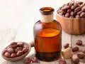 Butelka z bursztynowego szkła z płynnym złotym olejkiem jojoba na włosy, obok drewnianej łyżki z orzechami jojoba.