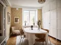 Comedor con mesa cubierta, sillas de madera y pared con papel tapiz dorado. Armarios blancos y suelo de parqué.