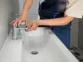Man con mono azul trabajando en un lavabo blanco, ajustando un grifo cromado.