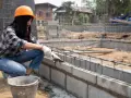 Une ouvrière en casque orange pose du ciment sur un mur en parpaing. Le chantier progresse.