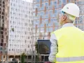 Ingeniero con casco y chaleco reflectante revisa un portátil frente a edificios en construcción. A pesar de un posible **ite desfavorable**, su mirada es decidida.