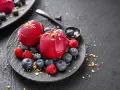 Zwei Kugeln Campari Sorbet mit frischen Beeren und Streuseln auf einem schwarzen Teller.