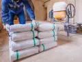 Budowlaniec układa worki z cementem, przygotowując się do mieszania betonu w proporcjach 1 do 3. W tle betoniarka i wiadra.