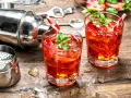 Zwei erfrischende Erdbeer-Cocktails mit Minze und Eiswürfeln, bereit zum Genießen. Ein Shaker und Bar-Werkzeuge deuten auf das kunstvolle Alkohol mischen hin.