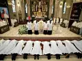 Klerycy w białych szatach leżą na posadzce kościoła, w geście pokory. Obok nich stoją inni duchowni, a w tle widać ołtarz i portret papieża.