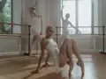 Trzy baletnice w białych strojach ćwiczą podnoszenia w tańcu. Jedna siedzi na podłodze, dwie inne stoją w pozycjach.