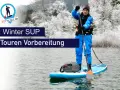 Person im blauen Neoprenanzug bereitet sich auf eine Winter SUP Tour vor. Die Landschaft ist mit Schnee bedeckt.