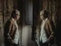 Uomo anziano con giacca marrone e camicia bianca, riflesso in uno specchio. L'arredamento suggerisce un'eleganza d'altri tempi, forse un omaggio a Balenciaga di dove è.