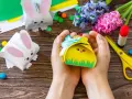 Ręce trzymają żółte pudełko w kształcie kurczaka, gotowe jako prezenty na Wielkanoc dla dzieci w szkole. Obok leżą papierowe zajączki i kolorowe cukierki.