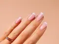 Dłoń z idealnym manicurem francuskim, paznokcie pomalowane na różowo z białymi końcówkami. Na serdecznym palcu złoty pierścionek z brylantem.