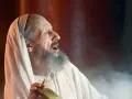 An elderly man, possibly Zechariah in the Bible, with a grey beard and wearing a white robe and head covering, cries out with his mouth open.