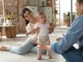 Rodzice zachęcają dziecko do pierwszych kroków. Uśmiechnięta mama i tata z otwartymi ramionami motywują malucha do chodzenia.