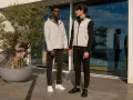 Due uomini con un moderno stile outfit uomo, uno con giacca grigia e jeans, l'altro con gilet grigio e pantaloni sportivi neri.