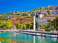 Malowniczy port w Chorwacji z łodziami przycumowanymi do nabrzeża. W tle widać wzgórze z zabudowaniami i zamek.
