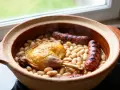 Un plat de cassoulet maison fumant dans une cocotte en terre cuite, avec un confit de canard doré et des saucisses grillées.