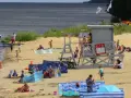Słoneczna puck plaża tętni życiem. Ludzie opalają się, kąpią i bawią na piasku. W tle widać wieżę ratowniczą i drzewa.