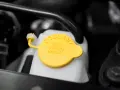 Close-up of a car's coolant reservoir with a yellow cap. The cap reads "COOLANT SEE OWNERS MANUAL," indicating where to find instructions on how to put coolant in car.
