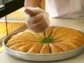 Ręka w rękawiczce posypuje zielonymi pistacjami okrągłą blachę z baklavą. To właśnie baklava co to za deser!