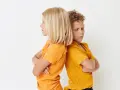Zwei Kinder stehen mit verschränkten Armen Rücken an Rücken. Ihre Mimik deutet auf **Gewalt unter Geschwistern** hin, ein Streit, der sie trennt.