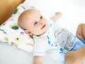Uśmiechnięty bobas leży na białej pościeli, opierając główkę na poduszce ortopedycznej dla niemowląt z motywem żółwi.
