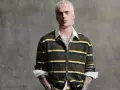 Joven con pelo rubio platino y tatuajes lleva un jersey de rayas verdes y amarillas, mostrando que **que jerseys se llevan este año** son los de estilo retro.