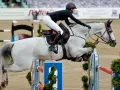 Reiterin mit weißem Pferd überwindet Hindernis. Ein Moment aus dem Springreiten, der die Dynamik der Klassen zeigt.
