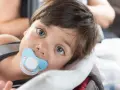 Małe dziecko z niebieskim smoczkiem Tommee Tippee w buzi, zapatrzone w dal.