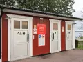 Röd byggnad med toaletter vid en sjö. Skyltar visar att camping är förbjuden. En trevlig rastplats med toalett för resenärer.