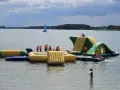 Dzieci w kamizelkach ratunkowych bawią się na dmuchanych wodnych atrakcjach. Idealne miejsce, gdzie na wakacje z dziećmi, z żaglówką na horyzoncie.