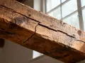 Gros plan sur une poutre en bois fissurée, témoin silencieux d'une maison qui craque toujours au même endroit. La lumière du jour filtre à travers une fenêtre floue.