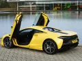 Żółty McLaren Artura z otwartymi drzwiami, stojący nad jeziorem przy nowoczesnym budynku.