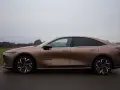 Brązowy samochód elektryczny Mazda6e widziany z boku na tle pochmurnego nieba i krajobrazu.