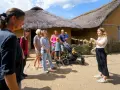 Familienurlaub in Haithabu: Eine Gruppe erkundet historische Häuser mit Strohdächern, während eine junge Frau eine Führung gibt.